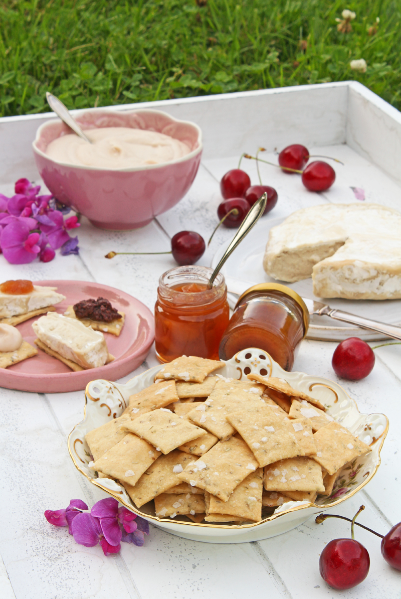 Picknick uppdukat på en vit träbricka, med en skål salta dinkelkex, en vitmögelost, en skål hummus och små marmeladburkar. På brickan ligger röda körsbär och rosa blommor utspridda.