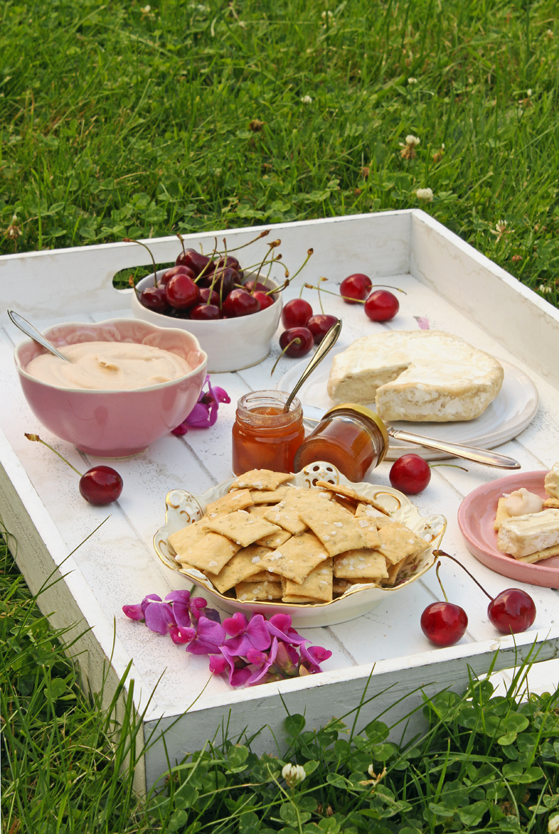 Picknick uppdukat på en vit träbricka, med en skål salta dinkelkex, en vitmögelost, en skål hummus, en skål körsbär och små marmeladburkar. På brickan ligger några körsbär och rosa blommor utspridda.