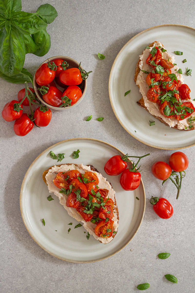Två tallrikar med surdegstoast med syrlig bönröra och rostade tomater, omgivna av färska tomater och basilikakvistar.