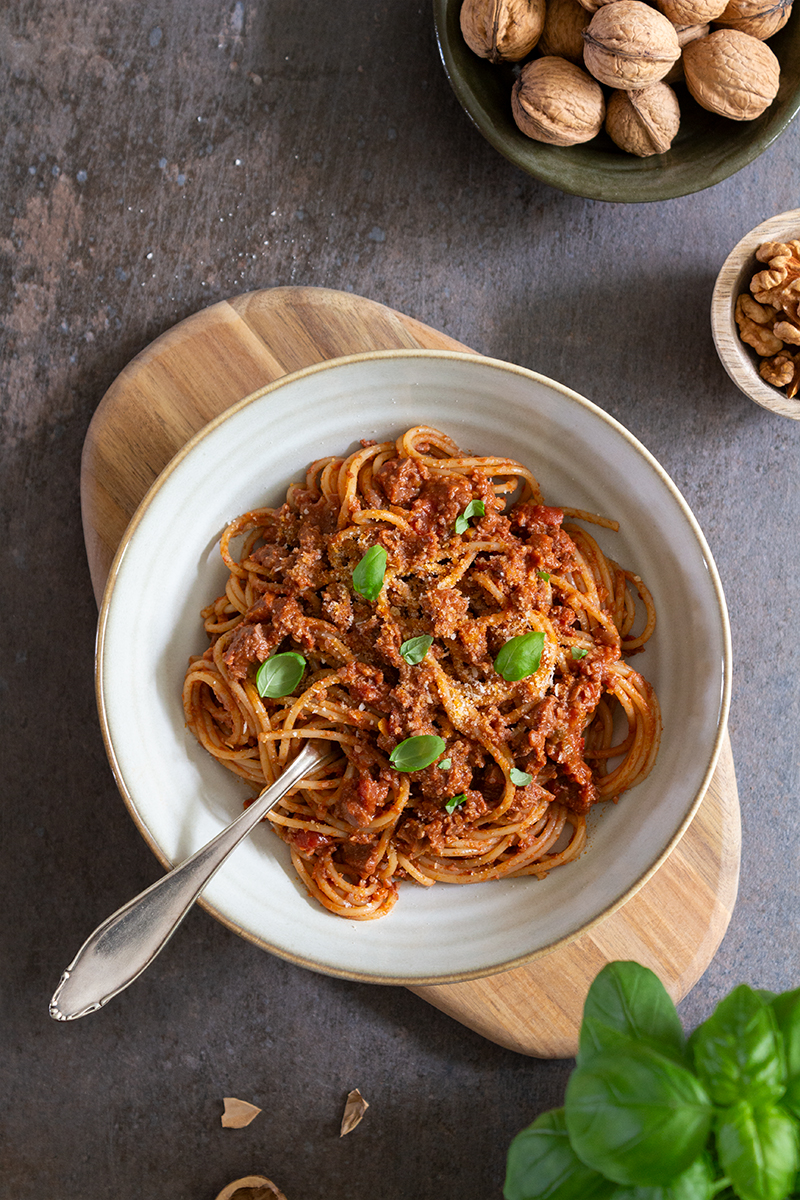En vit skål med tempeh och valnötsbolognese, stående på en träskärbräda. Bolognesen är toppad med basilikablad, och i pastan är en gaffel nedstucken. I kanterna skymtas skålar med valnötter samt basilikablad.
