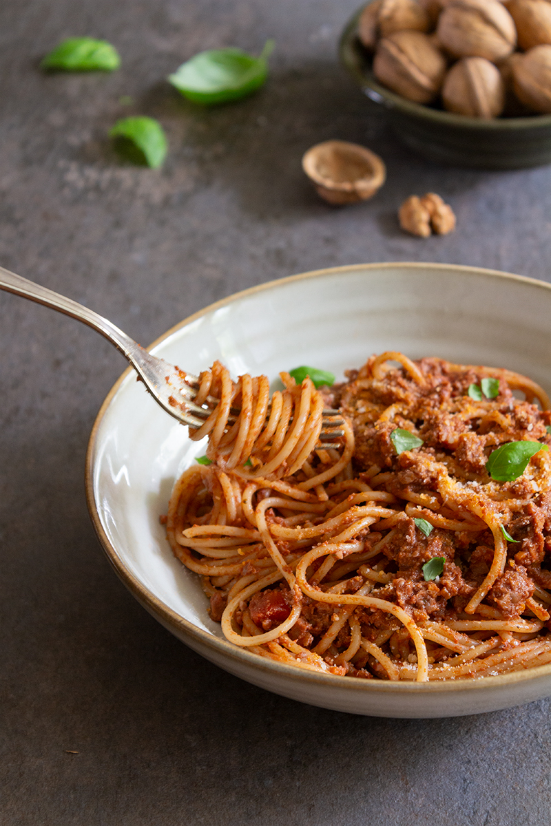 En skål med bolognese sedd från sidan. Bolognesen är toppad med basilikablad, och någon tar upp pasta ur skålen med en gaffel. I bakgrunden skymtas valnötter och basilika.
